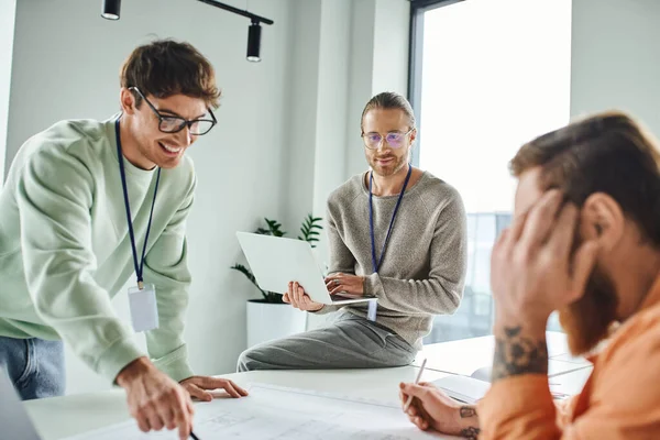 Optimistic designer in eyeglasses pointing at blueprint near colleague with laptop and tattooed businessman on blurred foreground, working on startup project, collaboration in architectural studio — Stock Photo