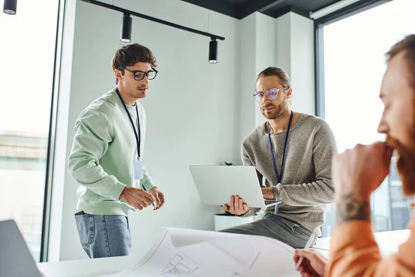 Thoughtful architectural designer in eyeglasses working on laptop near thoughtful business colleagues and blueprint on blurred foreground, creative coworking in modern office environment — Stock Photo