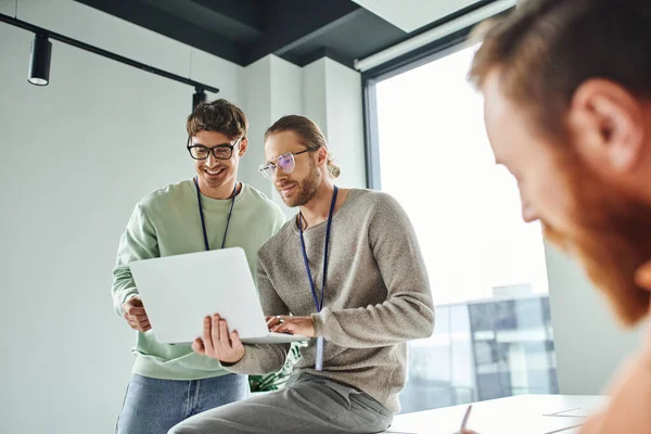 Positive emotion, successful and smiling designers in eyeglasses looking at laptop near colleague working on blurred foreground in modern coworking space of architectural studio — Stock Photo