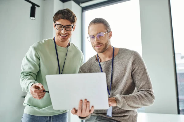 Lächelnder Architekturdesigner mit Brille, der auf Laptop zeigt, während er mit Kollegen in modernem Coworking-Büro an einem neuen Projekt arbeitet, Konzept der Kreativität und erfolgreicher Geschäftszusammenarbeit — Stockfoto