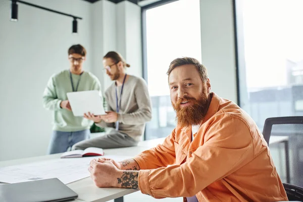 Optimistic, tattooed and bearded designer looking at camera near blueprint and colleagues working on new business project on blurred background in contemporary architectural studio — Stock Photo