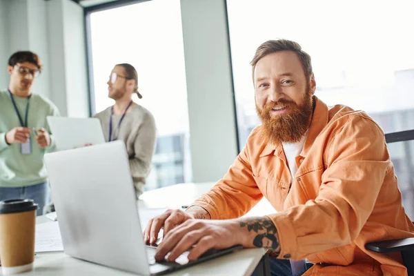 Cheerful bearded and tattooed architect working on laptop near takeaway drink in paper cup and looking at camera while colleagues talking on blurred background in coworking design studio — Stock Photo
