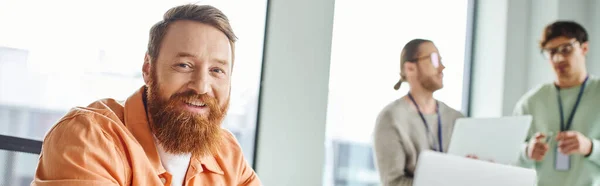 Portrait of cheerful and creative bearded architect looking at camera in modern coworking environment of design studio near colleagues with laptop on blurred background, banner — Stock Photo