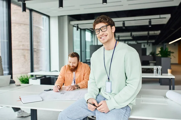Joyful entrepreneur in eyeglasses and casual clothes sitting on work desk and looking at camera while bearded architect working with blueprint in modern coworking space of design studio — Stock Photo