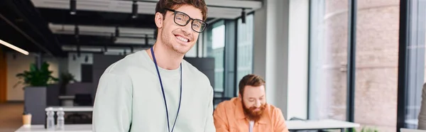 Overjoyed and successful businessman in eyeglasses looking at camera while bearded architect working on new project on blurred background in modern design studio, banner — Stock Photo