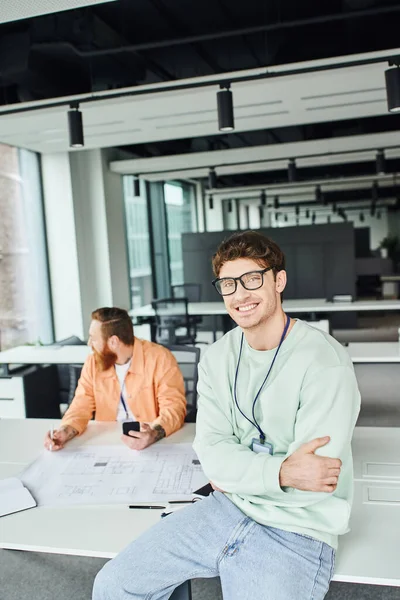 Satisfied entrepreneur in eyeglasses sitting on work desk with folded arms and smiling at camera next to bearded architect holding smartphone and working with blueprint on blurred background — Stock Photo