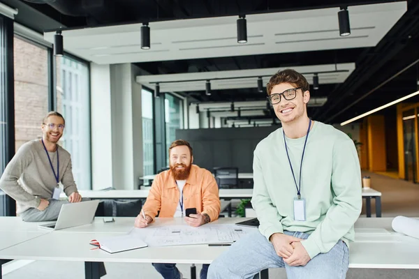 Happy team lead in eyeglasses looking at camera near smiling architects working on startup project with blueprint, laptop and mobile phone in contemporary design studio, successful teamwork concept — Stock Photo