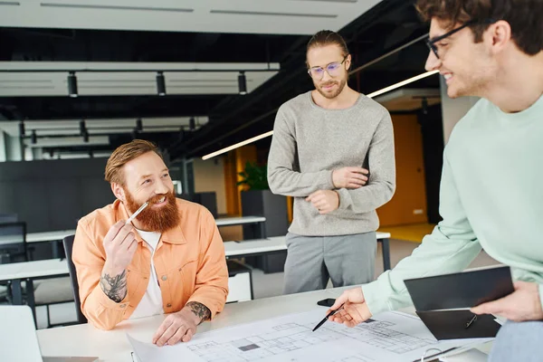 Architect in eyeglasses holding notebook and pointing with pen at blueprint near cheerful, bearded and tattooed designer in contemporary coworking office of architectural studio — Stock Photo