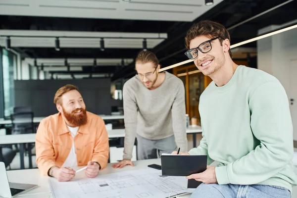 Pleased businessman in eyeglasses holding notebook and looking at camera next to creative architects working with blueprint near laptop in contemporary coworking space of design studio — Stock Photo