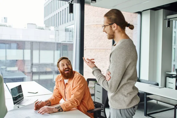 Entrepreneur in eyeglasses discussing new business project with bearded architectural designer laughing near blueprint and laptop in modern coworking office, creative collaboration concept — Stock Photo