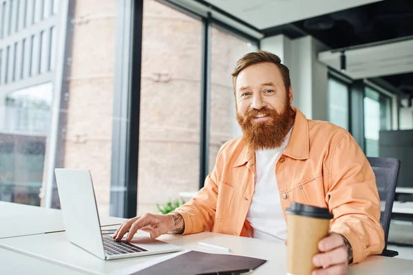 Положительные эмоции бородатый и татуированный человек с выпивкой на вынос сидя на рабочем месте рядом папка и ноутбук и улыбаясь на камеру в современной офисной среде, концепция успеха бизнеса — стоковое фото