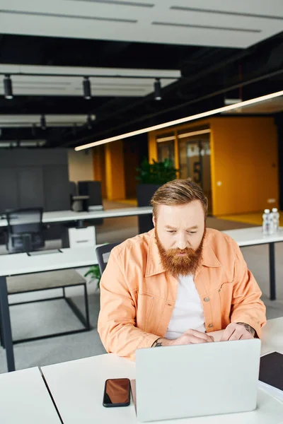 Fokussierter, bärtiger und tätowierter Geschäftsmann, der auf Laptop schaut, während er am modernen Arbeitsplatz in der Nähe von Smartphones mit leerem Bildschirm in modernen Büroräumen sitzt und an einem Startup-Projekt arbeitet — Stockfoto