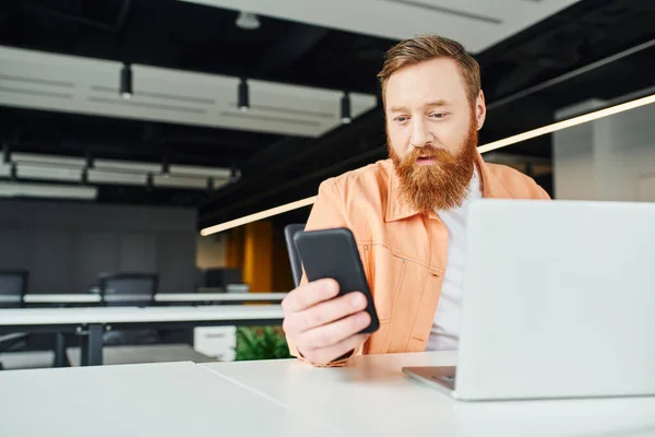 Бородатий і стильний бізнесмен в повсякденному одязі, дивлячись на мобільний телефон, сидячи на робочому місці біля ноутбука і працюючи над стартап-проектом в коворкінгу сучасного офісу — стокове фото