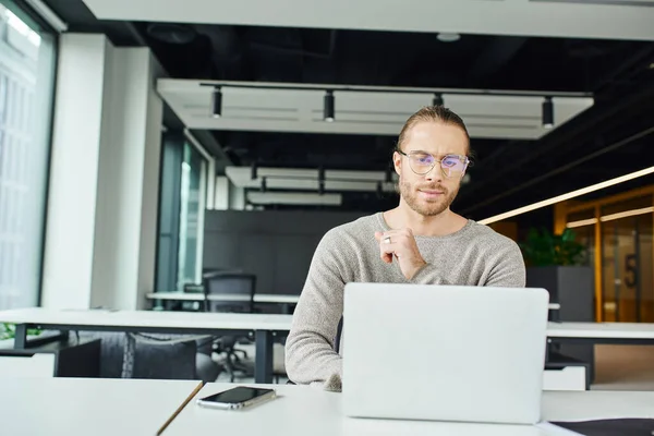 Erfolgreicher Unternehmer mit stylischer Brille, der auf den Laptop schaut, während er an einem Geschäftsprojekt in der Nähe von Mobiltelefonen mit leerem Bildschirm am Arbeitsplatz in der modernen Umgebung des Coworking Office arbeitet — Stockfoto