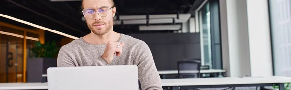 Вдумливий і концентрований бізнесмен в повсякденному одязі і стильних окулярах дивиться на ноутбук під час роботи над стартап-проектом в сучасному коворкінгу, банер з копіювальним простором — стокове фото