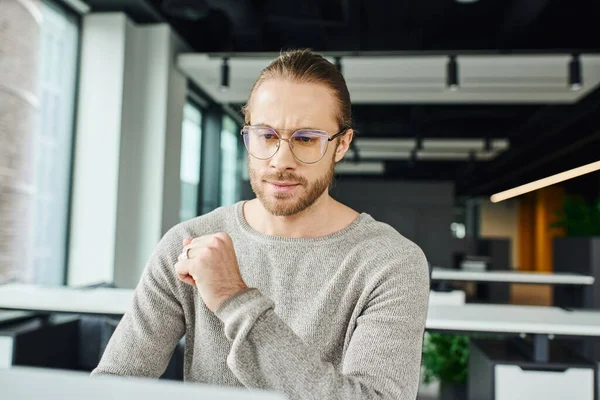 Серьезный бизнесмен в повседневной одежде и стильных очках, работающий над планированием стартапов, глядя на ноутбук и мышление в современном офисе, бизнес-успех и концепция производительности — стоковое фото