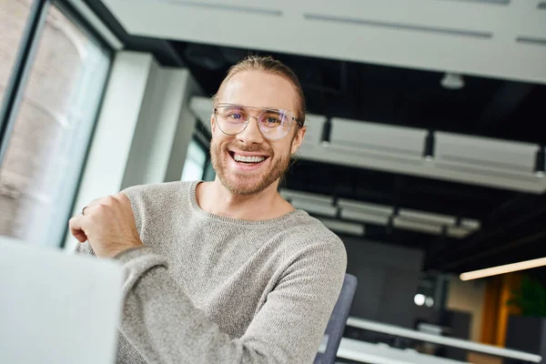 Профессиональный снимок восторженного бизнесмена в стильной одежде и очках, улыбающегося в камеру возле ноутбука на размытом переднем плане в современном офисе, бизнес-продуктивности и концепции успеха — стоковое фото