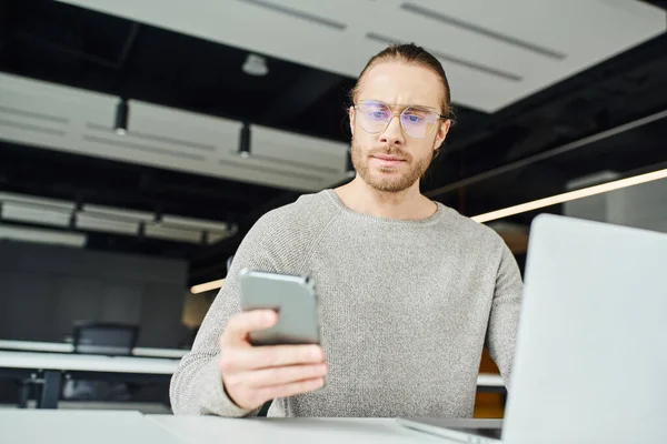 Unternehmer mit Brille und lässiger Kleidung, mit ernstem Gesichtsausdruck, der während der Arbeit an einem Startup-Projekt in der Nähe von Laptop auf verschwommenem Vordergrund in modernen Büroräumen aufs Handy schaut — Stockfoto