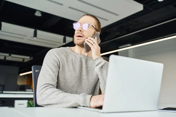 Thoughtful businessman in eyeglasses looking away and talking on mobile phone while sitting near laptop on work desk, startup project planning in contemporary office space — Stock Photo