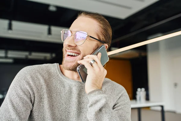 Emprendedor emocionado en gafas con estilo y ropa casual mirando hacia otro lado y riendo durante la conversación en el teléfono móvil en la oficina moderna, productividad del negocio y el concepto de éxito - foto de stock