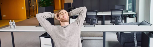 Tired businessman in stylish eyeglasses and casual clothes sitting with closed eyes and hands behind head while resting at workplace in modern office environment, banner — Stock Photo