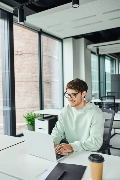 Cheerful entrepreneur in stylish eyeglasses and casual clothes sitting at modern workplace and networking on laptop near folder and coffee to go in contemporary office, successful business concept — Stock Photo