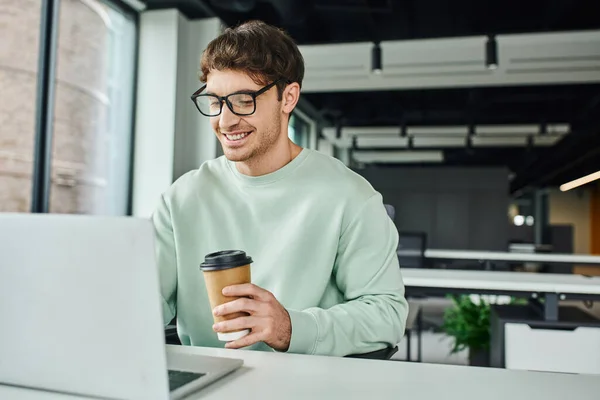 Улыбающийся бизнесмен в повседневной одежде и стильных очках держит бумажный стаканчик с напитком на вынос и работает на ноутбуке в современной офисной среде, успешная бизнес-концепция — стоковое фото