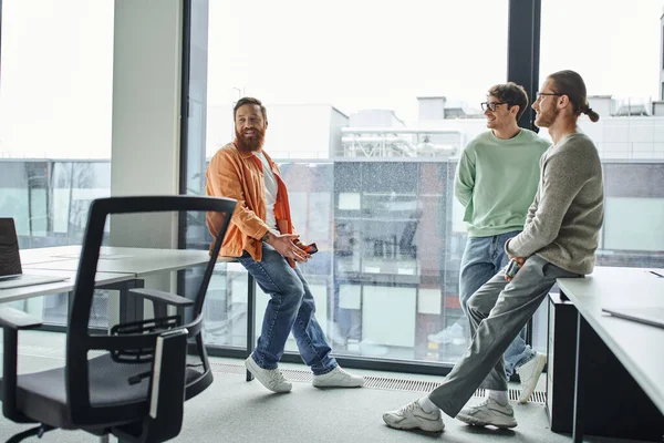 Empresarios positivos en ropa casual elegante sonriendo mientras están de pie cerca de escritorios de trabajo y grandes ventanas en el entorno de coworking moderno, concepto de colaboración exitosa - foto de stock