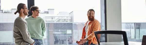 Бодрый бородатый бизнесмен держит мобильный телефон и смотрит в сторону улыбающихся коллег в современном коворкинг-офисе с большими окнами, концепция успешного сотрудничества, баннер — стоковое фото