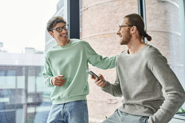 Joyful entrepreneur in eyeglasses and casual clothes touching shoulder of business partner sitting with smartphone and hand in pocket near large office window, concept of successful collaboration — Stock Photo