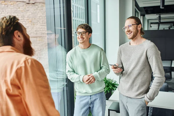 Smiling businessmen in eyeglasses and casual clothes listening to bearded team lead talking on blurred foreground near large windows in contemporary office, concept of successful collaboration — Stock Photo