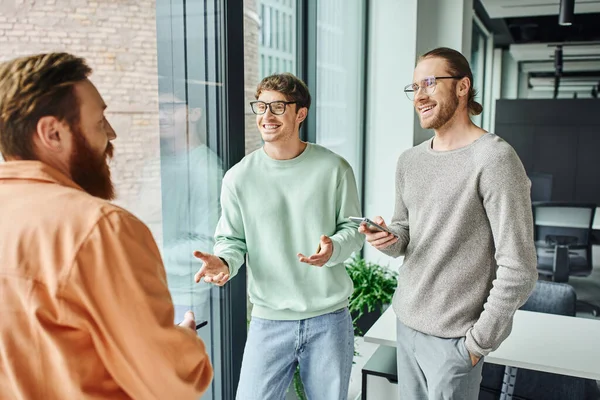 Homens de negócios sorridentes em óculos gesticulando ao discutir projeto de startup com empreendedor em primeiro plano borrado perto de grandes janelas no escritório moderno, conceito de colaboração bem sucedida — Fotografia de Stock
