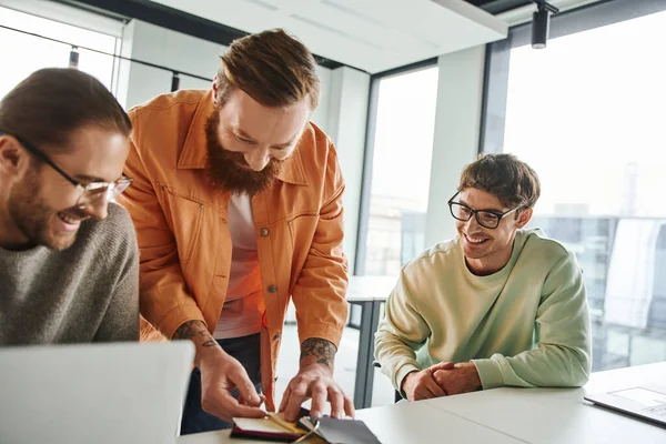 Bearded and tattooed designer showing color samples to cheerful and stylish entrepreneurs in eyeglasses sitting near laptops in contemporary coworking environment, teamwork and business creativity — Stock Photo