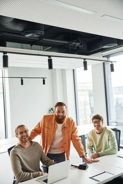 Professionelle Aufnahme erfolgreicher Unternehmer in stilvoller Freizeitkleidung, die in die Kamera lächeln, in der Nähe von Laptop und Farbmustern auf dem Schreibtisch im zeitgenössischen Designstudio, kreatives Business-Team — Stockfoto