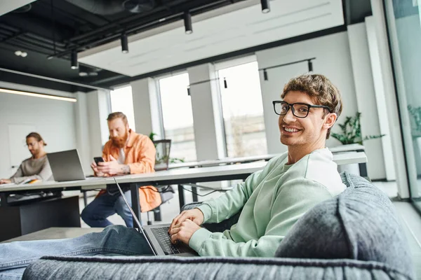 Joyful team lead in eyeglasses sitting on couch with laptop, planning startup and looking at camera near business team working on blurred background, productive coworking concept — Stock Photo