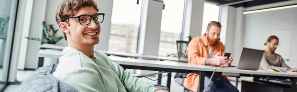 Erfolgreicher und optimistischer Teamleiter mit stylischer Brille, der in einem modernen Büroumfeld in der Nähe von Kollegen mit verschwommenem Hintergrund in die Kamera lächelt, produktives Coworking-Konzept, Banner — Stockfoto