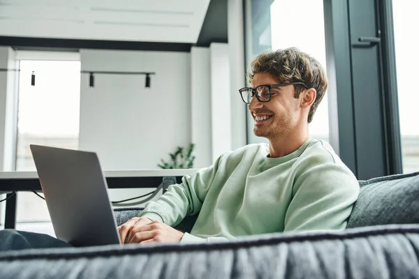 Cheerful and successful businessman in stylish eyeglasses and casual clothes sitting on comfortable couch in office lounge of coworking space and networking on laptop, business inspiration concept — Stock Photo