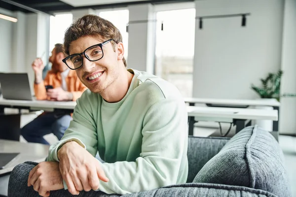 Happy entrepreneur in stylish eyeglasses and casual clothes sitting in armchair and smiling at camera near colleague working on blurred background in modern office, productive coworking concept — Stock Photo