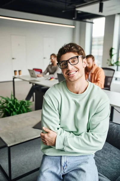 Empresário alegre em óculos e roupas casuais sentado com os braços dobrados e olhando para a câmera, enquanto os colegas que trabalham perto de laptops em fundo turvo, conceito de coworking produtivo — Fotografia de Stock