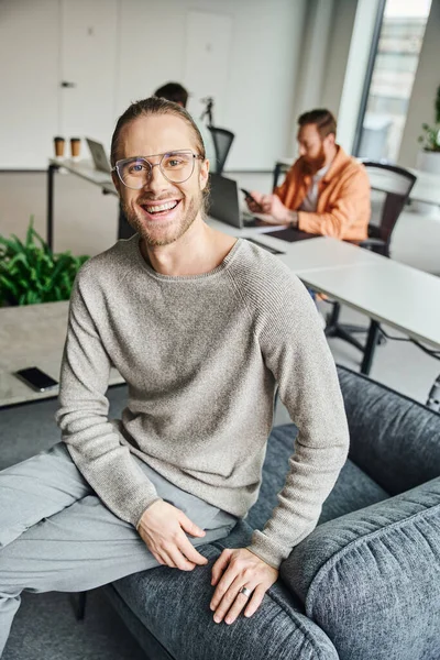 Empreendedor feliz em óculos elegantes olhando para a câmera no ambiente de co-trabalho moderno perto de colegas que trabalham em fundo turvo, conceito de empreendedorismo bem sucedido — Fotografia de Stock