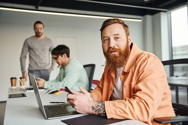 Bearded and tattooed businessman in stylish casual clothes holding smartphone and looking at camera near laptop and colleagues working on blurred background in contemporary coworking space — Stock Photo