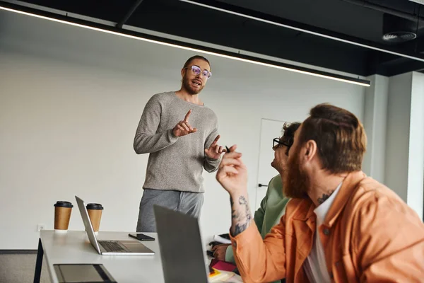 Entrepreneur in eyeglasses and casual clothes talking and showing attention gesture to business partners working near laptops and paper cups in modern coworking environment — Stock Photo