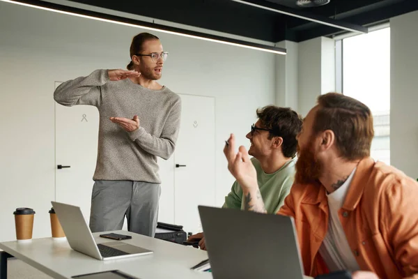 Team lead in eyeglasses showing quantity gesture while discussing startup with excited colleagues sitting at laptops in contemporary coworking office, business collaboration and teamwork concept — Stock Photo