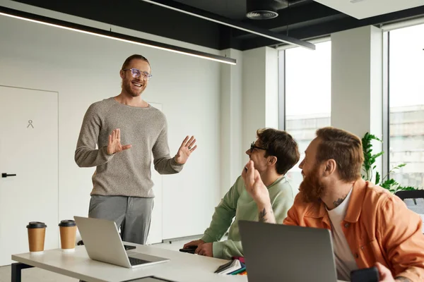 Fröhlicher Geschäftsmann in lässiger Kleidung und stylischer Brille mit Stop-Geste neben Kollegen, Laptops und Pappbechern im modernen Büro, erfolgreiches Teamwork-Konzept — Stockfoto