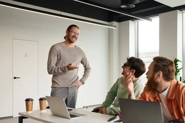 Positiver Unternehmer mit stylischer Brille, der mit der Hand auf der Hüfte steht und mit dem Finger zeigt, während er mit seinem Team im modernen Coworking-Büro an Laptops sitzend Geschäftsprojekte diskutiert — Stockfoto