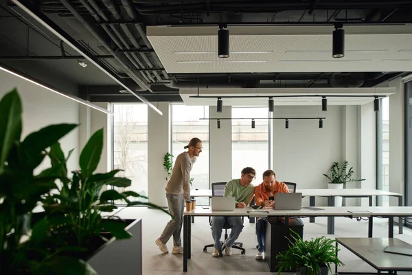 Творческая и успешная команда в стильной повседневной одежде, работающая рядом с ноутбуками на рабочем месте в современной коворкинговой среде с высокотехнологичным интерьером, концепцией делового сотрудничества, баннером — стоковое фото