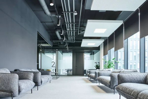 Espaçosa área de espera com sofás cinzentos e confortáveis, grandes janelas e plantas verdes no ambiente de escritório contemporâneo, conceito de organização do espaço de trabalho — Fotografia de Stock