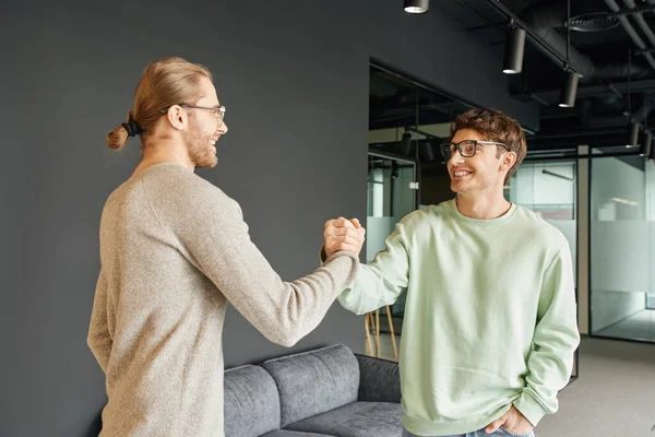 Cheerful and stylish entrepreneurs in eyeglasses and casual clothes shaking hands and confirming agreement in lounge of modern coworking office, successful collaboration concept — Stock Photo