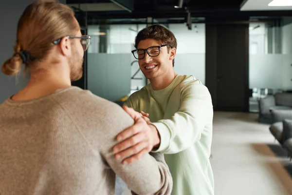 Happy businessman in casual clothes and eyeglasses shaking hands and touching shoulder of colleague while confirming deal in lounge of modern coworking space, successful partnership concept — Stock Photo