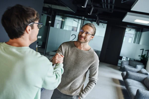Pleased businessman in eyeglasses and stylish casual clothes shaking hands with partner while closing deal in lounge of contemporary coworking office, successful collaboration concept — Stock Photo